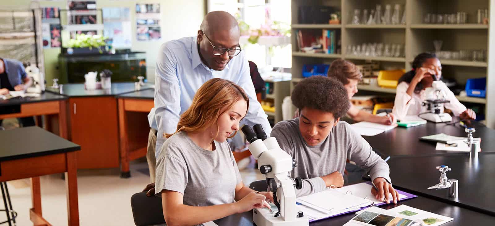 High School Students With Tutor Using Microscope In Biology Class